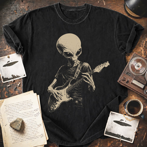 A black graphic t-shirt featuring a detailed beige print of an alien playing an electric guitar, styled in a flat-lay arrangement on a rustic wooden table with vintage props including a record player, coffee cup, and old papers.