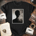 A black graphic t-shirt laid flat on a rustic wooden table, featuring a central square print of a smoking human silhouette in grayscale. The shirt is styled with vintage props including a cassette tape, a coffee cup, an open book, and small UFO-themed cards.