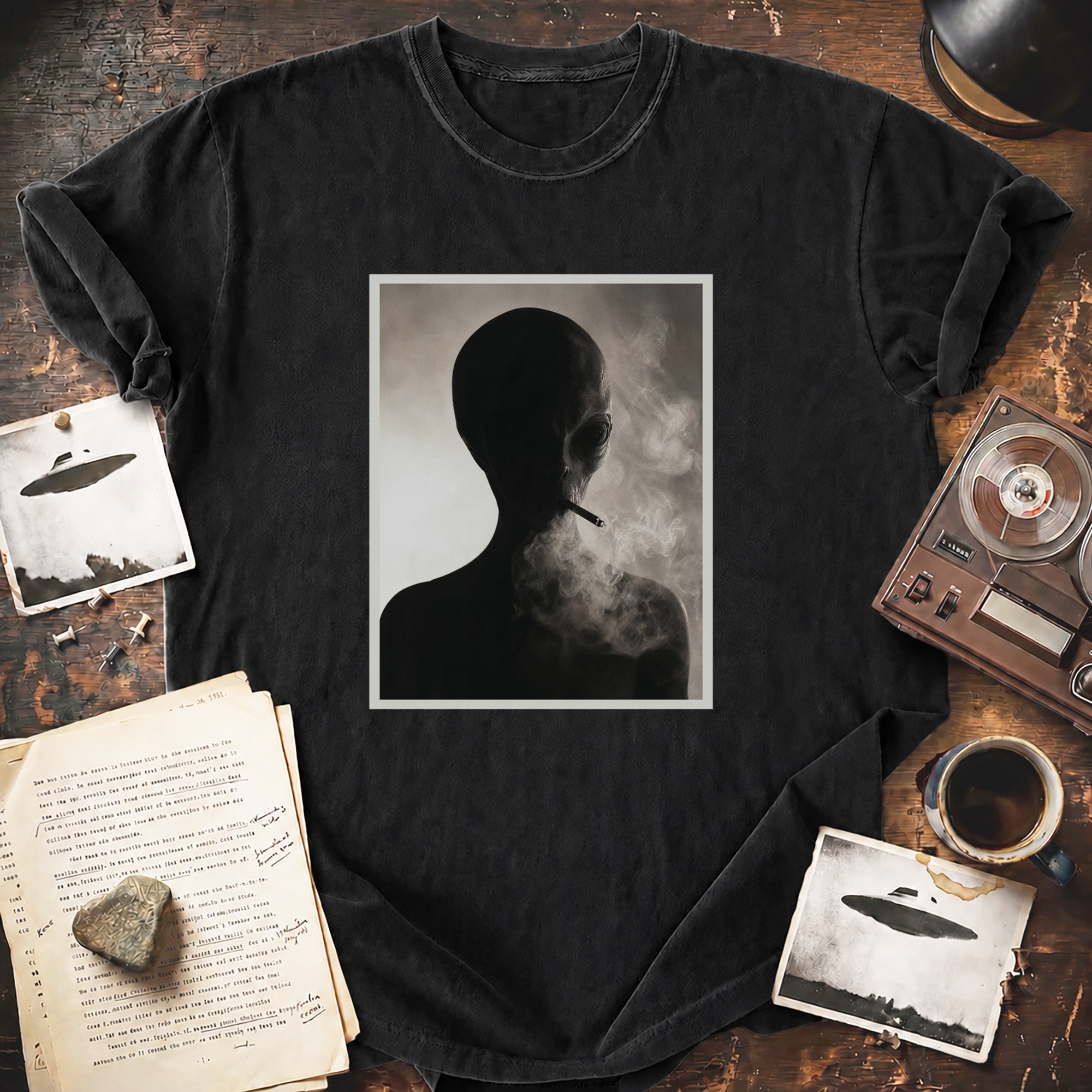 A black graphic t-shirt laid flat on a rustic wooden table, featuring a central square print of a smoking human silhouette in grayscale. The shirt is styled with vintage props including a cassette tape, a coffee cup, an open book, and small UFO-themed cards.
