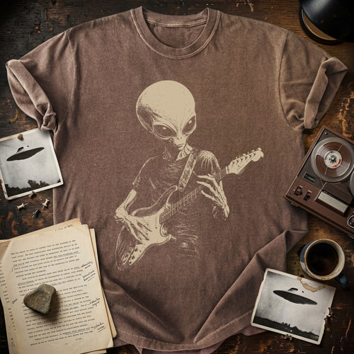 A brown short-sleeve t-shirt with a beige graphic of an alien playing an electric guitar, styled in a flat-lay on a dark wooden table with vintage props including a tape recorder, coffee cup, open book, and UFO-themed photos.