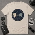A cream-colored short-sleeve t-shirt with a circular graphic print featuring a space-themed design with two profiles (one beige, one gray) and the text 'INTERGALACTIC FEDERATION' and 'CONSCIOUSNESS EXCHANGE'. The shirt is displayed in a flat-lay arrangement on a gray textured background, surrounded by lifestyle props including a notebook, sunglasses, a polaroid camera, and a small card.
