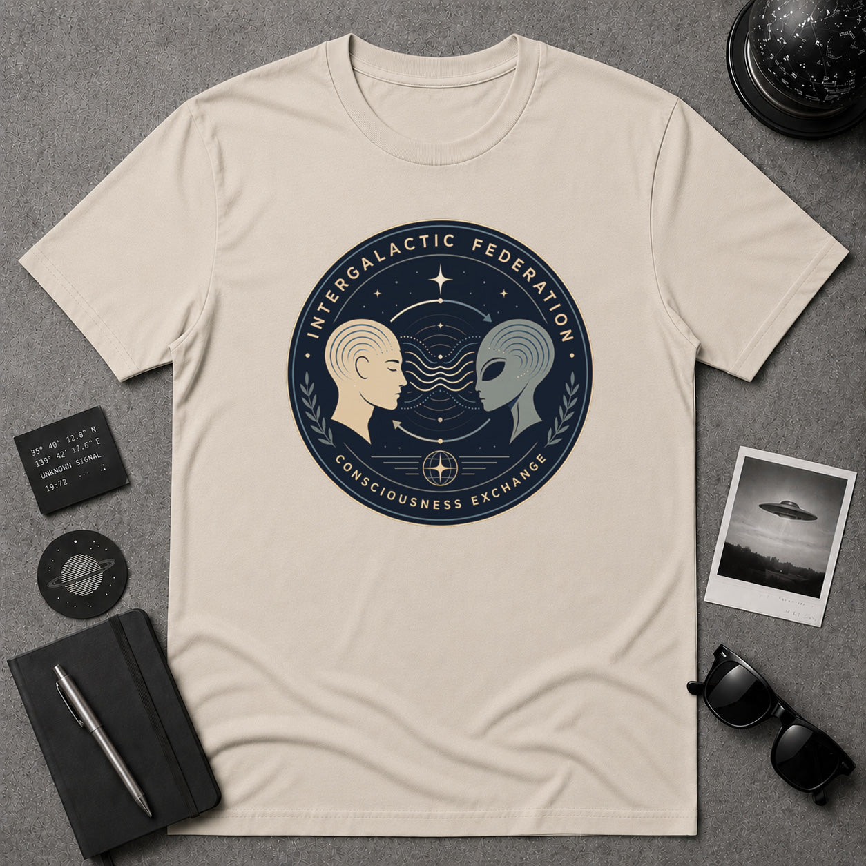 A cream-colored short-sleeve t-shirt with a circular graphic print featuring a space-themed design with two profiles (one beige, one gray) and the text 'INTERGALACTIC FEDERATION' and 'CONSCIOUSNESS EXCHANGE'. The shirt is displayed in a flat-lay arrangement on a gray textured background, surrounded by lifestyle props including a notebook, sunglasses, a polaroid camera, and a small card.