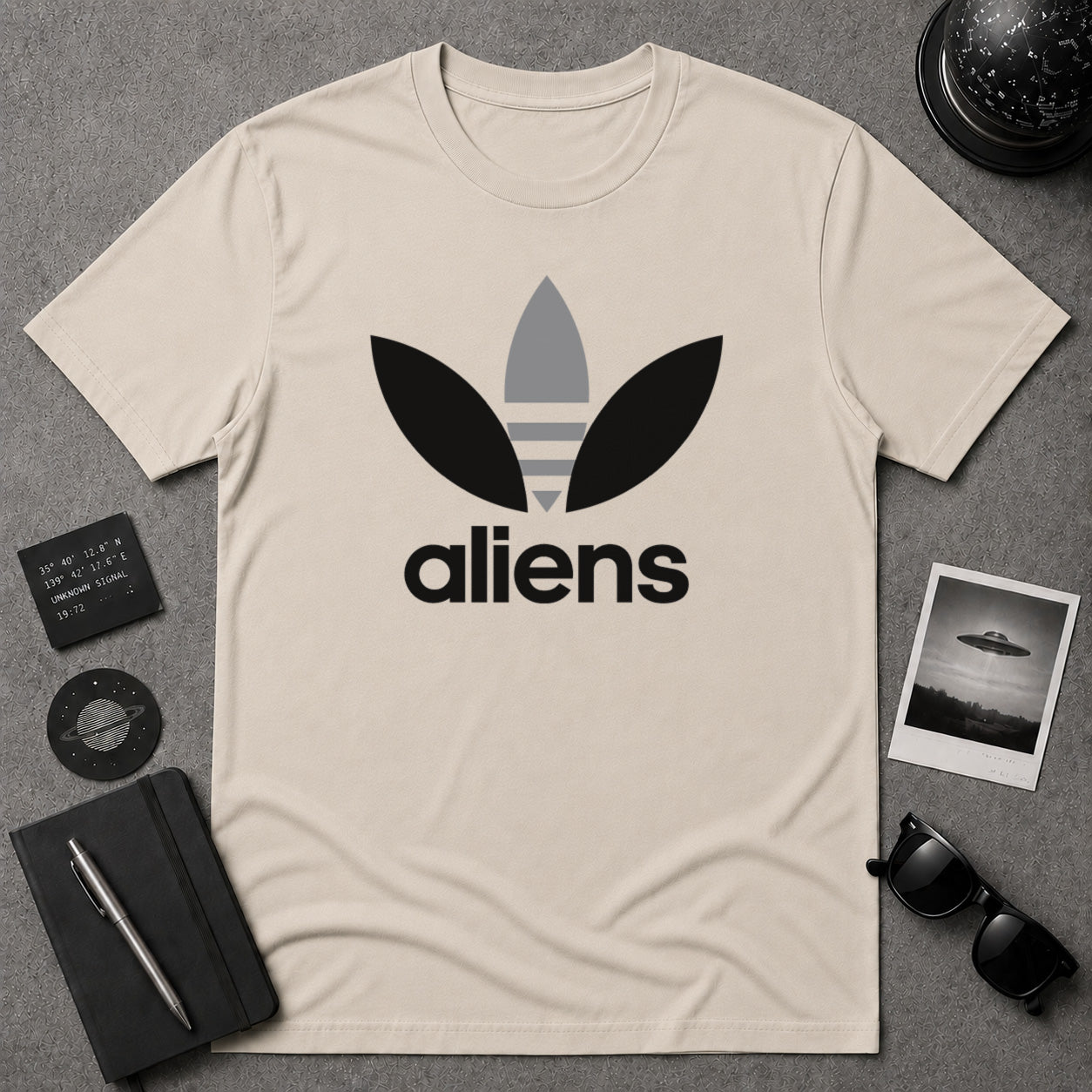 A beige graphic t-shirt featuring a black and gray alien-themed logo and the word 'aliens' printed on the front, visually reminiscent of the classic Adidas logo. This is a parody shirt.